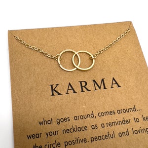 Könnte beinhalten: Eine goldfarbene Halskette mit zwei ineinandergreifenden Kreisen, präsentiert auf einer braunen Karte. Auf der Karte steht das Wort "KARMA" in großen Buchstaben, sowie der Text "what goes around, comes around..." und weitere inspirierende Sätze.