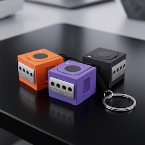 May include: Three miniature GameCube console keyrings in orange, purple, and black. Each cube-shaped keyring features a black top button, silver port details, and a small lanyard. One keyring has a silver ring attached.