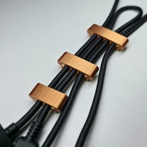 Puede incluir: Primer plano de cables negros sujetos por tres organizadores de cables de color cobre. Los organizadores son rectangulares y parecen ser de metal. Los cables están agrupados y ordenados.