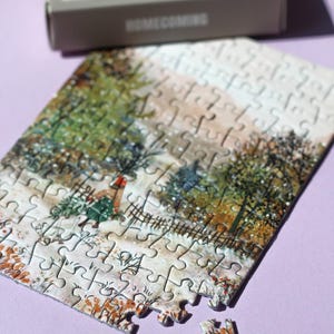 May include: A partially assembled jigsaw puzzle depicting a winter scene with a person carrying a Christmas tree. The puzzle pieces are scattered on a purple surface, with the box in the background. The box reads "HOMECOMING".
