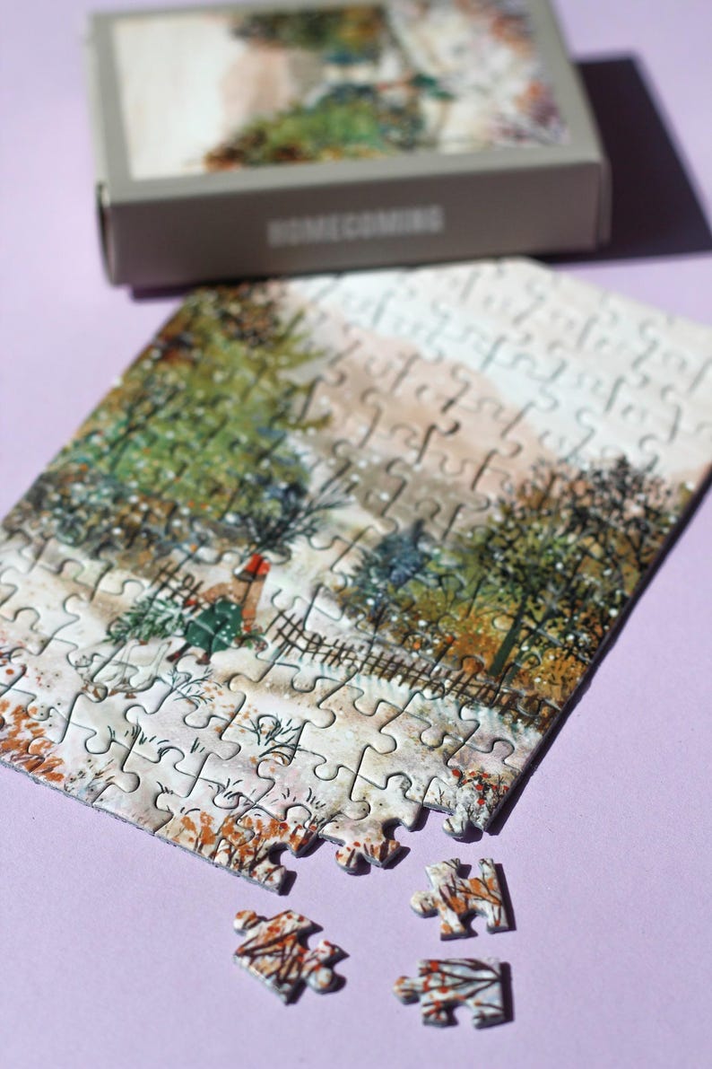 May include: A partially assembled jigsaw puzzle depicting a winter scene with a person carrying a Christmas tree. The puzzle pieces are scattered on a purple surface, with the box in the background. The box reads "HOMECOMING".