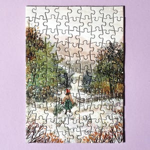 May include: A completed jigsaw puzzle showing a winter landscape. The image features a person walking through snow, carrying branches. The puzzle includes a fence, trees, and a muted colour palette of greens, browns, and whites.