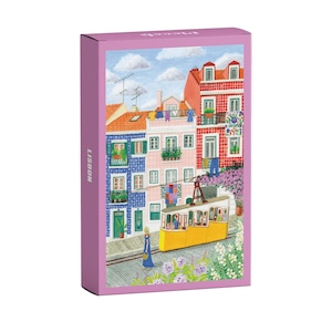 Könnte beinhalten: Eine rechteckige Schachtel mit einer Illustration von Lissabon. Das Bild zeigt eine gelbe Straßenbahn, Gebäude in Rosa, Rot und Blau sowie eine Person in einem blauen Kleid. Das Wort "LISBON" ist auf der Seite der Schachtel aufgedruckt.
