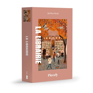 Könnte beinhalten: Ein 500-teiliges Puzzle mit rosa Hintergrund. Das Puzzle zeigt eine detaillierte Illustration eines Buchladens mit Menschen und Hunden draußen. Die Worte "LA LIBRAIRIE" sind vertikal auf der Seite und der Vorderseite der Box aufgedruckt.