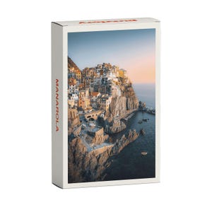 Könnte beinhalten: Eine farbenfrohe Stadtlandschaft, die auf einer Klippe über dem Meer gebaut wurde. Die Gebäude sind ein Mix aus Pastellfarben und das Wasser ist tiefblau. Der Himmel ist in sanftem Rosa und Orange gehalten, was auf einen Sonnenuntergang hindeutet. Der Text "MANAROLA" ist vertikal auf der linken Seite des Bildes gedruckt.