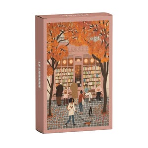 Puede incluir: Una caja rectangular rosa con una ilustración de una librería. La imagen muestra a personas frente a una tienda con la palabra "LIBRAIRIE" sobre la puerta. La caja también tiene las palabras "LA LIBRAIRIE" impresas en el lateral.