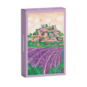 Könnte beinhalten: Eine lila Schachtel mit einem Gemälde eines provenzalischen Dorfes. Das Bild zeigt eine Stadt am Hang mit Gebäuden und Bäumen, ein Lavendelfeld und den Text "LA PROVENCE" an der Seite.