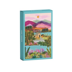 Könnte beinhalten: Eine farbenfrohe, illustrierte Puzzlebox mit einer malerischen Aussicht auf einen See mit einer kleinen Kirche, Bergen und Blumen. Die Box ist blau und hat die Aufschrift "Lake Tekapo" auf der Seite.