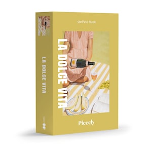 Könnte beinhalten: Ein 500-teiliges Puzzle mit einer Frau in einem rosa Kleid, die auf einer gestreiften Decke mit einem Glas Wein und einer Flasche Champagner sitzt. Das Puzzle trägt den Titel "La Dolce Vita" und wird von Piecely hergestellt.