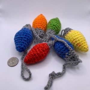 May include: A string of six crocheted Christmas light bulbs in various colors: blue, orange, green, red, and yellow. Each bulb is connected by a gray crocheted cord. The lights are handmade and appear to be soft.
