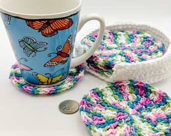 May include: Set of crocheted coasters in white, pink, green, and blue. The coasters are round and have a textured design. A mug with a butterfly pattern is sitting on one of the coasters.