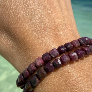 Puede incluir: Dos pulseras de piedras preciosas de color rojo oscuro. Las cuentas son de corte cuadrado y tienen un aspecto facetado. Las pulseras se llevan en una muñeca, con un fondo borroso del océano.