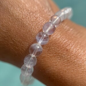 May include: A clear, beaded bracelet with a light purple hue. The bracelet is made of round, translucent beads strung together on a clear elastic cord. The bracelet is worn on a wrist, with a turquoise background.