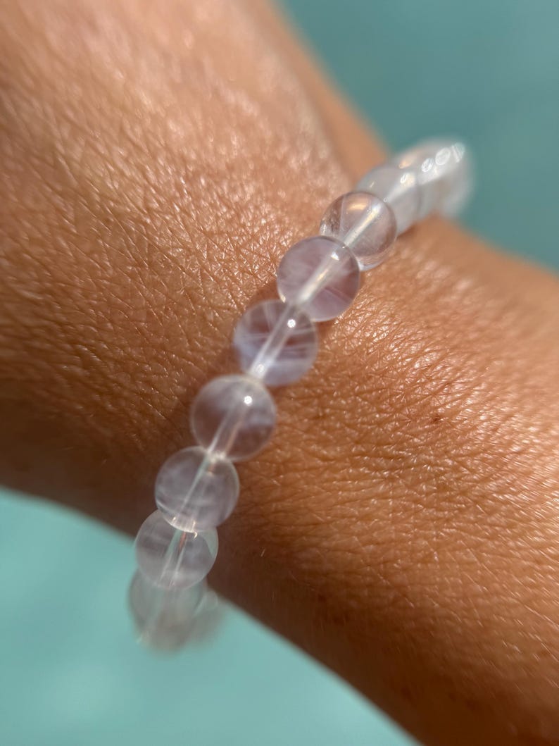 May include: A clear, beaded bracelet with a light purple hue. The bracelet is made of round, translucent beads strung together on a clear elastic cord. The bracelet is worn on a wrist, with a turquoise background.