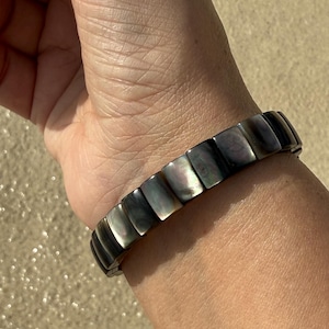 Puede incluir: Un brazalete de concha negra e iridiscente con forma rectangular. El brazalete está hecho de múltiples piezas pequeñas conectadas.