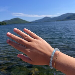 May include: A hand wearing a light blue gemstone bracelet and a gold chain bracelet against a backdrop of a lake and mountains.