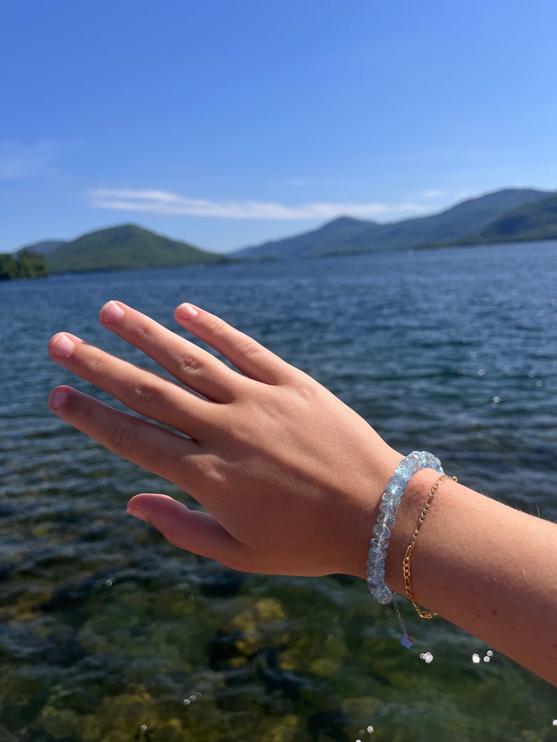May include: A hand wearing a light blue gemstone bracelet and a gold chain bracelet against a backdrop of a lake and mountains.