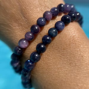 May include: Two beaded bracelets with dark blue and purple beads. The bracelets are made of small, round beads strung together. The beads have a polished, glossy finish. The bracelets are worn on a wrist.