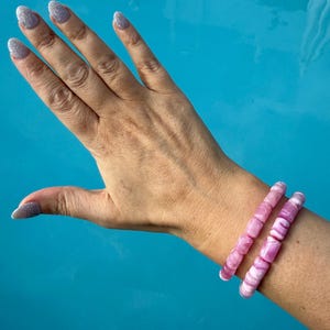 May include: Two pink and white beaded bracelets are worn on a wrist. The bracelets are made of cylindrical beads. The background is a bright blue, possibly water. The person's hand is visible with glittery nail polish.