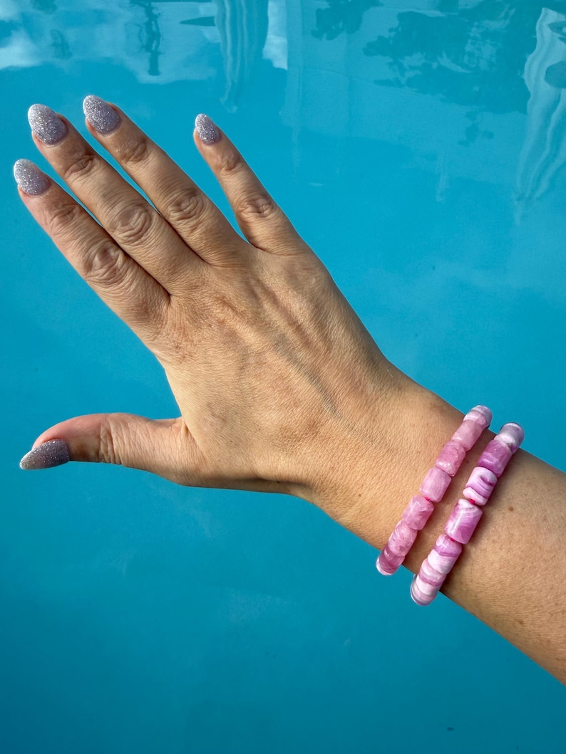 May include: Two pink and white beaded bracelets are worn on a wrist. The bracelets are made of cylindrical beads. The background is a bright blue, possibly water. The person's hand is visible with glittery nail polish.