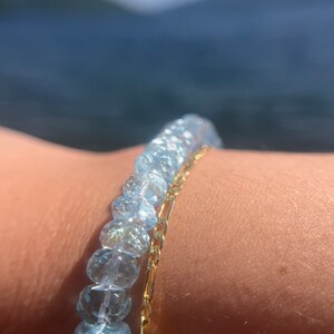 May include: A bracelet made of light blue gemstone beads and a gold chain. The bracelet is worn on a person's wrist.