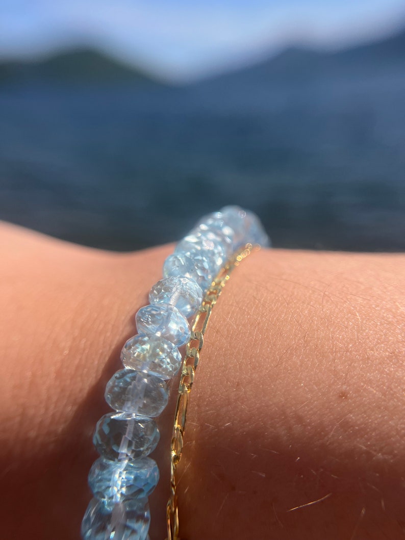 May include: A bracelet made of light blue gemstone beads and a gold chain. The bracelet is worn on a person's wrist.