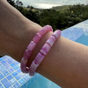 May include: Two pink beaded bracelets. The bracelets are made of rectangular beads in varying shades of pink and white. The bracelets are worn on a wrist, with a background of a pool and a scenic view of trees and sky.