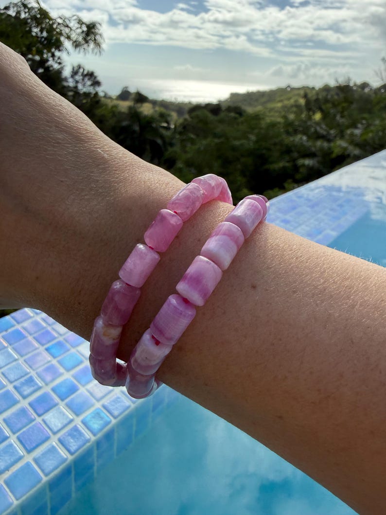 May include: Two pink beaded bracelets. The bracelets are made of rectangular beads in varying shades of pink and white. The bracelets are worn on a wrist, with a background of a pool and a scenic view of trees and sky.