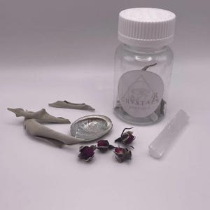 May include: A clear glass bottle with a white lid and a label that reads "CRYSTALS BOUTIQUE" is surrounded by sage bundles, a polished abalone shell, dried rosebuds, and a white selenite wand. The items are arranged on a white surface.