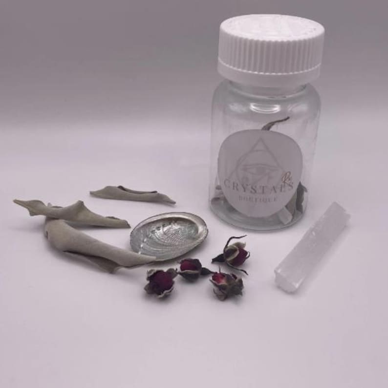 May include: A clear glass bottle with a white lid and a label that reads "CRYSTALS BOUTIQUE" is surrounded by sage bundles, a polished abalone shell, dried rosebuds, and a white selenite wand. The items are arranged on a white surface.
