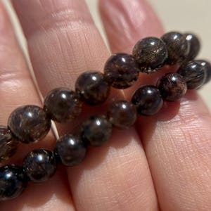 May include: Two strands of dark brown, round gemstone beads. The beads have a translucent quality with internal inclusions, creating a shimmering effect. The bracelet is held in a hand, showcasing the beads' polished surface and natural variations.