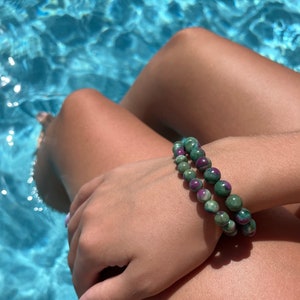 Może przedstawiać: Bransoletka z zielonych i fioletowych kamieni szlachetnych z okrągłymi koralikami. Bransoletka jest noszona na nadgarstku.