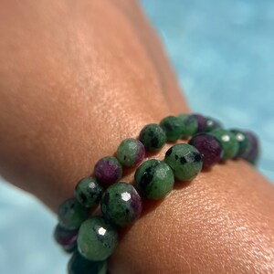 May include: A green and purple gemstone bracelet with faceted beads. The bracelet is worn on a person's wrist.