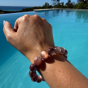 Peut inclure: Un bracelet de perles avec de grosses perles rondes, violettes et roses. Le bracelet est porté au poignet, avec une piscine bleue et l'océan en arrière-plan. Les perles sont translucides.