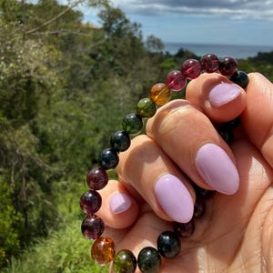 May include: A multi-colored beaded bracelet featuring a variety of gemstone beads in shades of red, green, orange, and black. The bracelet is held up against a natural outdoor background with green foliage and a view of the ocean.