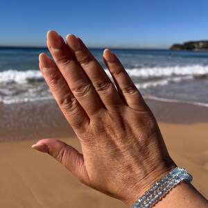 May include: A hand wearing a bracelet made of light blue aquamarine beads. The bracelet is made of three strands of beads and is worn on the wrist.