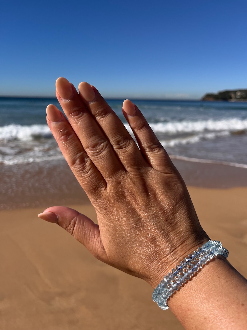 May include: A hand wearing a bracelet made of light blue aquamarine beads. The bracelet is made of three strands of beads and is worn on the wrist.