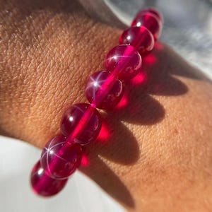 May include: A close-up of a bracelet featuring large, round, deep red beads. Each bead has a star-like effect, with lines of light radiating from the center. The bracelet is strung on a clear elastic band, and is worn on a wrist.