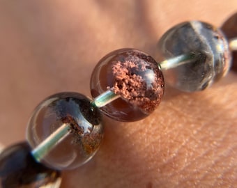 Puede incluir: Primer plano de un brazalete hecho de cuentas de cuarzo transparente con inclusiones marrones y negras. Las cuentas son redondas y tienen un acabado liso y pulido.