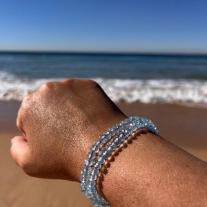 May include: A light blue aquamarine gemstone bracelet with multiple strands of beads. The bracelet is worn on a person's wrist against a background of a sandy beach and ocean.