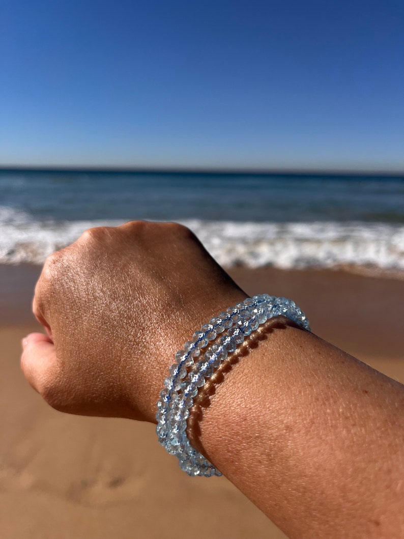 May include: A light blue aquamarine gemstone bracelet with multiple strands of beads. The bracelet is worn on a person's wrist against a background of a sandy beach and ocean.