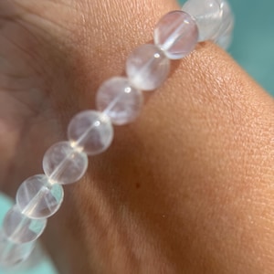 May include: A clear, beaded bracelet with a subtle, pearlescent sheen. The round beads are strung together, creating a delicate and elegant accessory. The bracelet is shown on a wrist, with a soft, neutral skin tone.