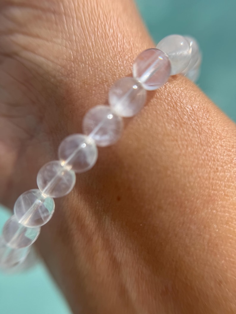 May include: A clear, beaded bracelet with a subtle, pearlescent sheen. The round beads are strung together, creating a delicate and elegant accessory. The bracelet is shown on a wrist, with a soft, neutral skin tone.
