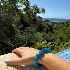 Puede incluir: Una pulsera de cuentas azules se lleva en la muñeca. La pulsera está hecha de cuentas grandes, redondas y translúcidas. El fondo muestra un exuberante paisaje verde con árboles y una vista del océano bajo un cielo azul claro.