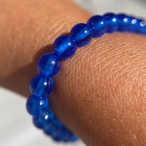 May include: A blue beaded bracelet with a shiny, iridescent finish. The bracelet is made of round beads and is worn on a person's wrist.