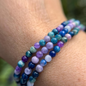 May include: A stack of four bracelets made with blue, green, and purple gemstone beads. The bracelets are layered on a person's wrist.