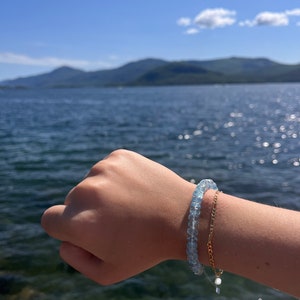 May include: A light blue gemstone bracelet with a gold chain bracelet on the same wrist. The bracelet is worn by a person with their hand outstretched against a background of a blue lake and mountains.