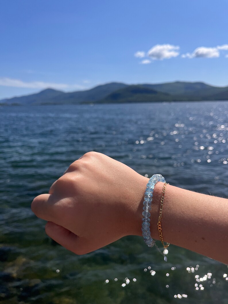 May include: A light blue gemstone bracelet with a gold chain bracelet on the same wrist. The bracelet is worn by a person with their hand outstretched against a background of a blue lake and mountains.