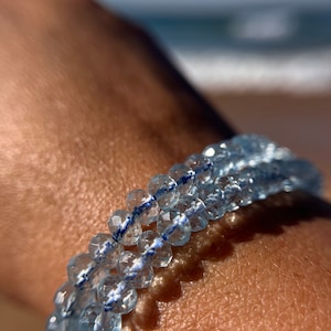 May include: A light blue gemstone bracelet with faceted beads and a silver thread running through the center.