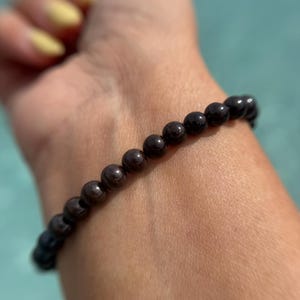 Pode incluir: Uma pulseira de contas castanhas escuras. A pulseira é feita de pequenas contas redondas enfiadas juntas. As contas têm um acabamento polido e refletem a luz. A pulseira é usada no pulso.